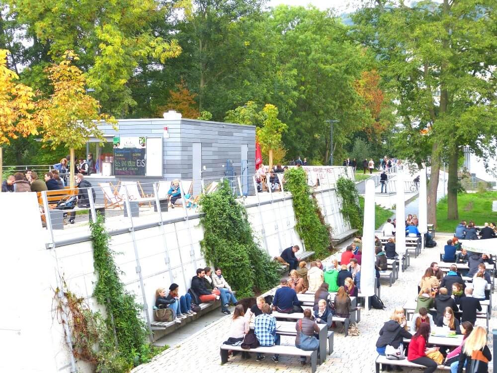 Blick auf die AUSZEIT und viele junge menschen die davor sitzen und essen und trinken