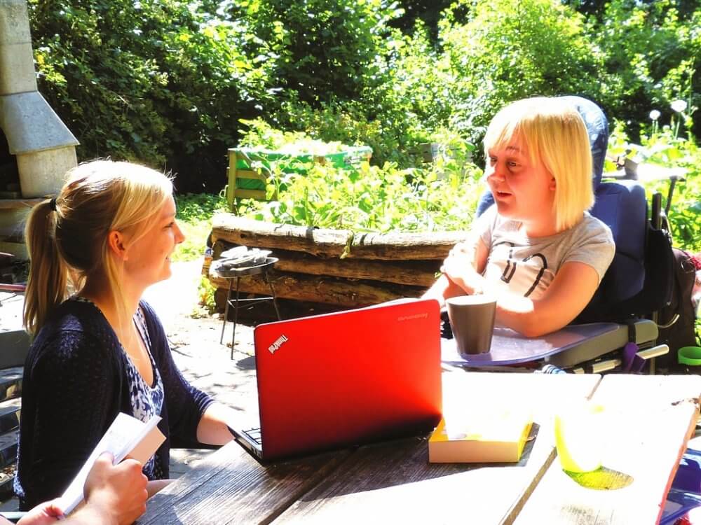 zwei junge Frauen sitzen am Tisch i Garten vor einem Laptop, eine Frau nutzt einen Rollstuhl